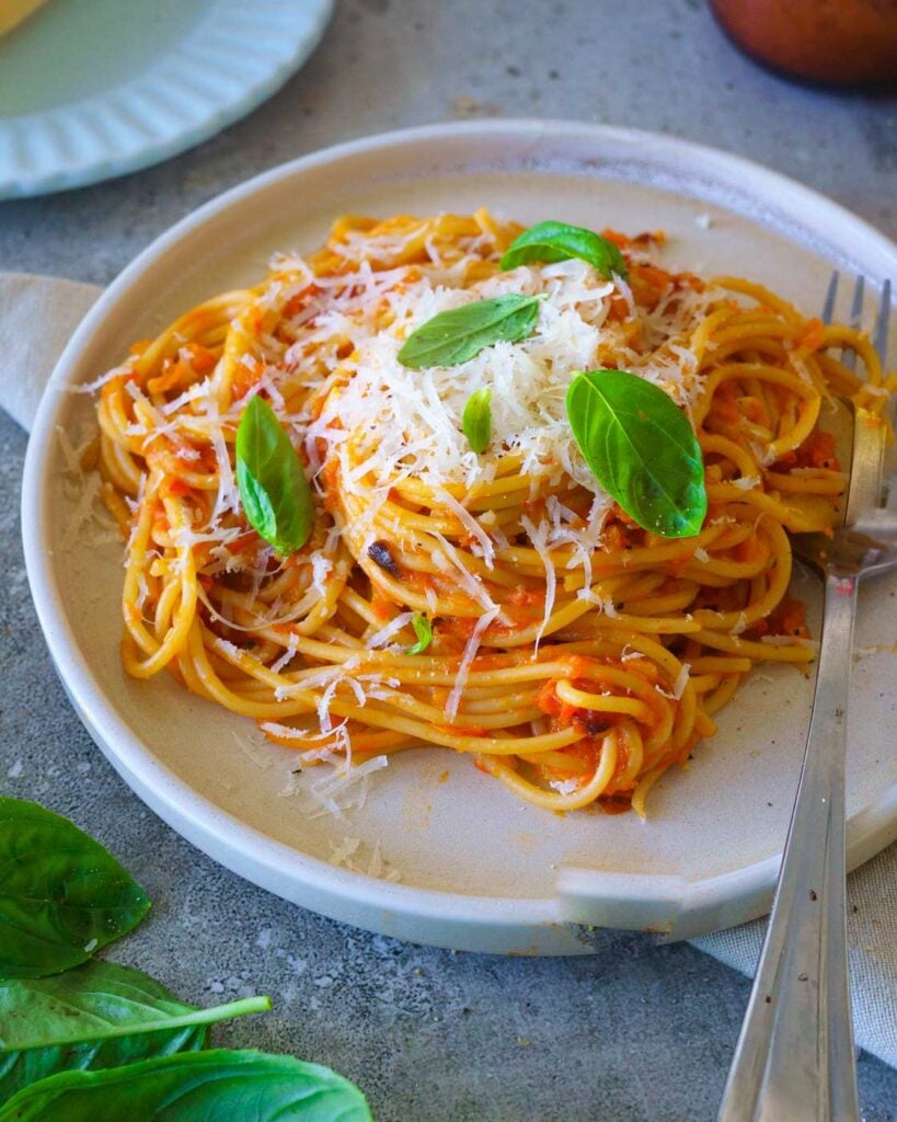 spaghetti tossed in roasted tomato and red pepper sauce on a white plate with basil leaves and parmesan