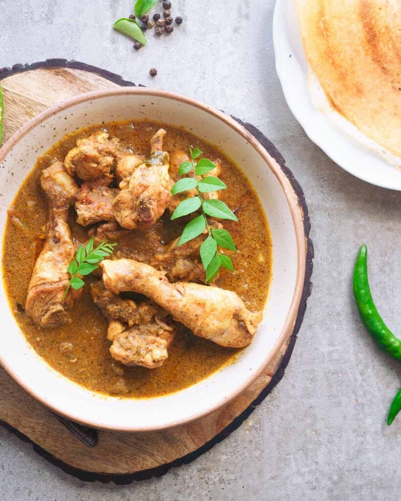 Pepper Chicken Curry inside a white shallow bowl on top of a wooden cutting board along with dosas and chilies