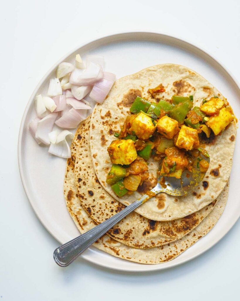 Paneer capsicum sabzi on a roti, on top of a beige plate with raw cut onions on the side.