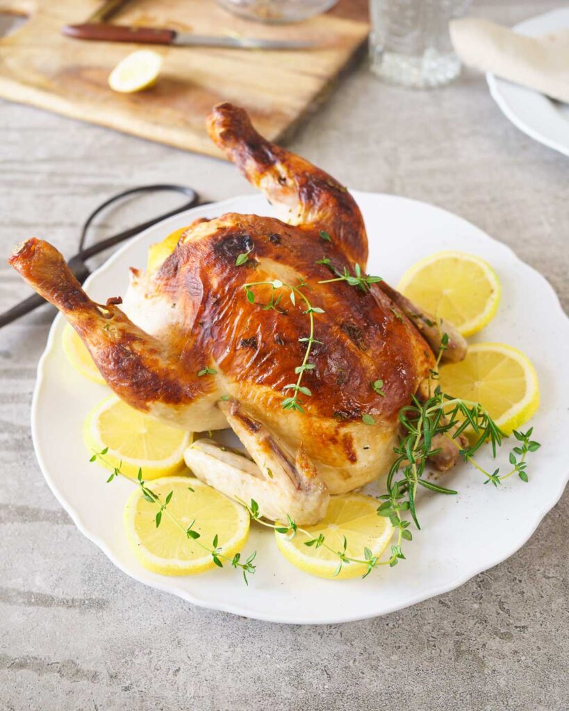 Buttermilk brined Roast chicken on top of a white plate served with lemon slices and fresh herbs.
