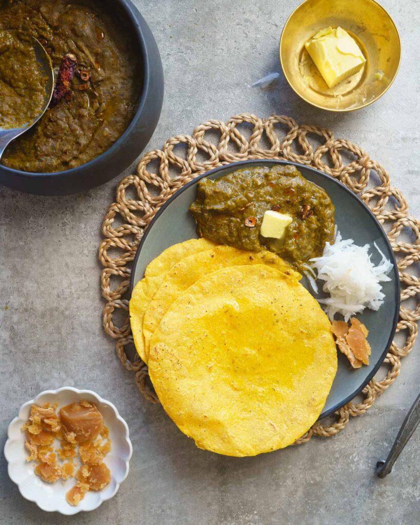 Sarson ka saag in a grey plate with a tempering of garlic, chili and ghee, topped with a knob of butter.SErved along with 3 makki ki roti or maize rotis.
Served along with grated radish & gur or jaggery