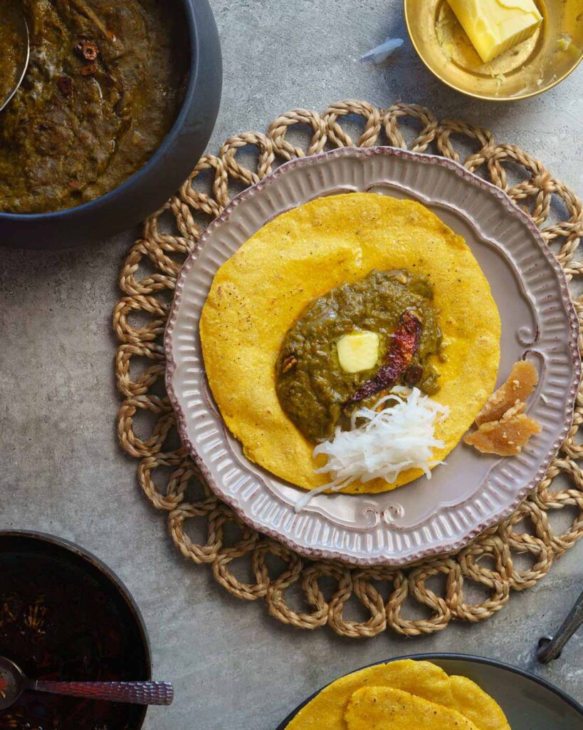Sarson ka saag in a brown plate with a tempering of garlic, chili and ghee, topped with a knob of butter.Served on top of  makki ki roti or maize roti along with grated radish & gur or jaggery