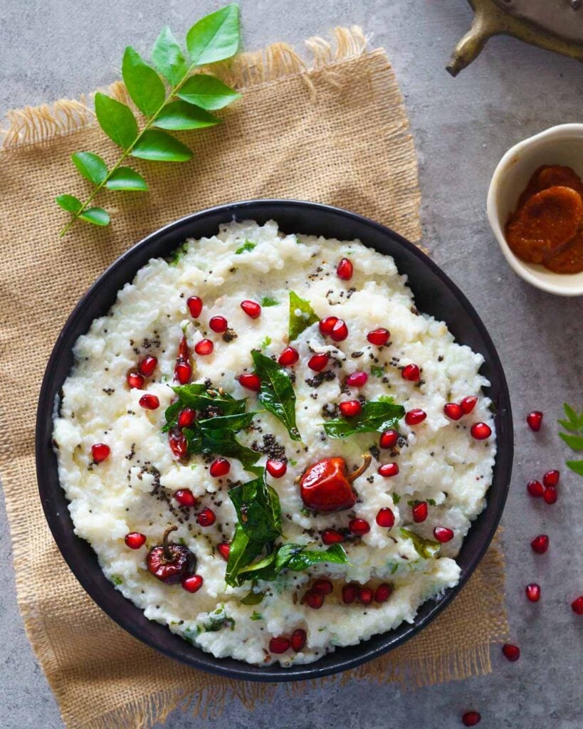 Curd Rice with pomegranate seeds, tempering of mustard seeds, curry leaves and chilies on a grey background , served with lime pickle.