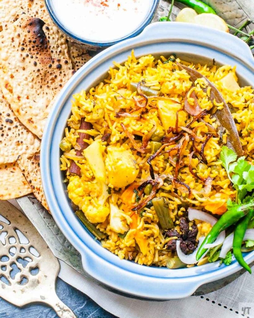 Instant Pot Vegetable Biryani with fried onions on top, in a blue earthen bowl served with papad, raita and raw onions