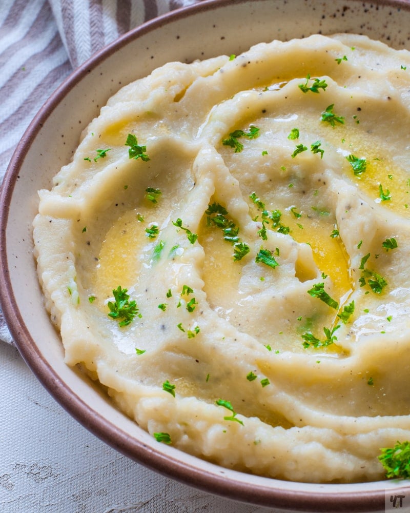 Mashed Potatoes made in the instant pot in a white shallow bowl with a brown rim, garnished with parsley and a  knob of butter