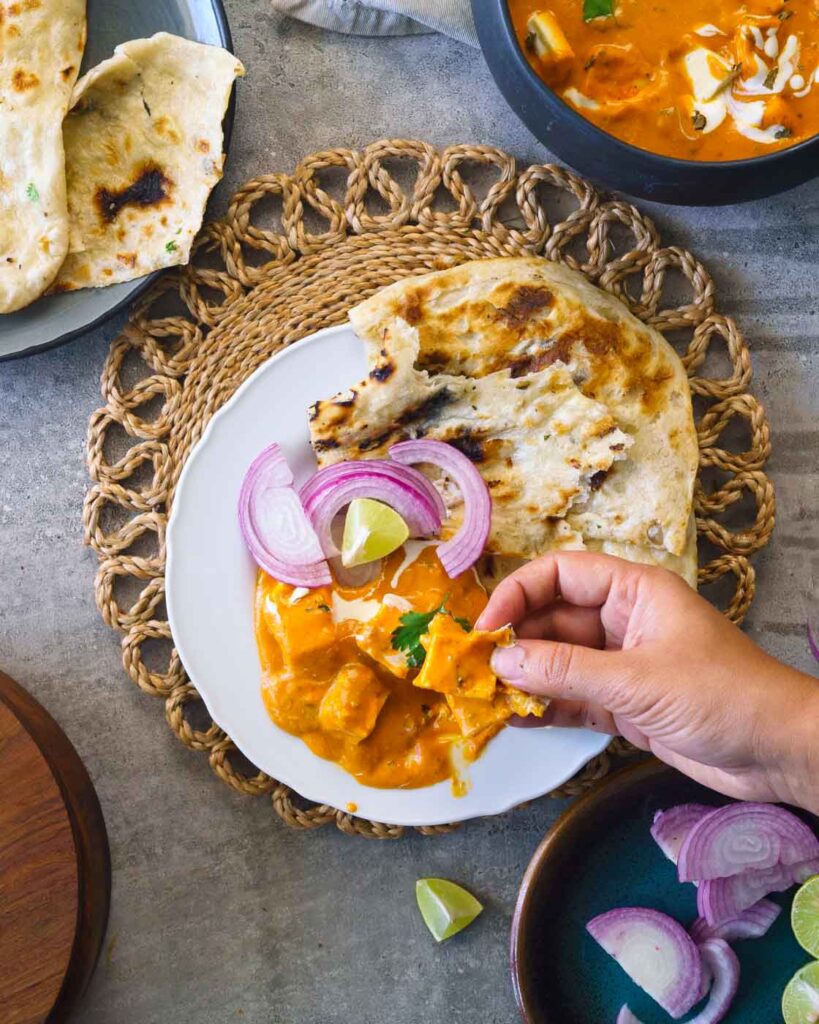 paneer butter masala on a white plate with naan, onions and lemon