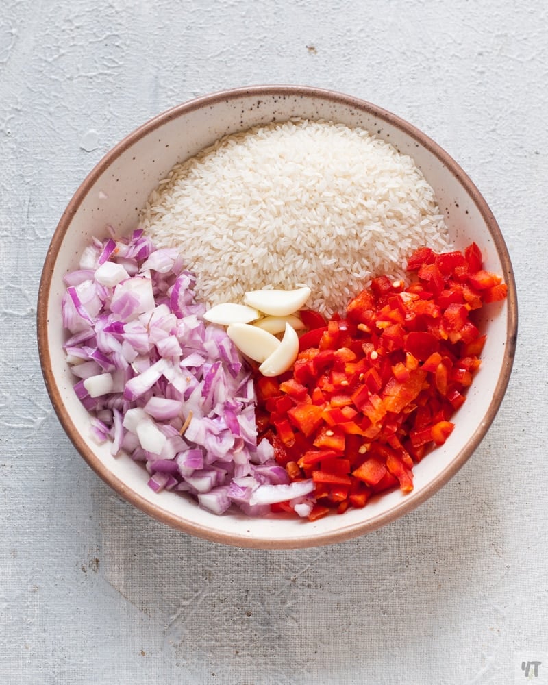 Chopped onion, garlic, red bell pepper and rice in a white bowl.