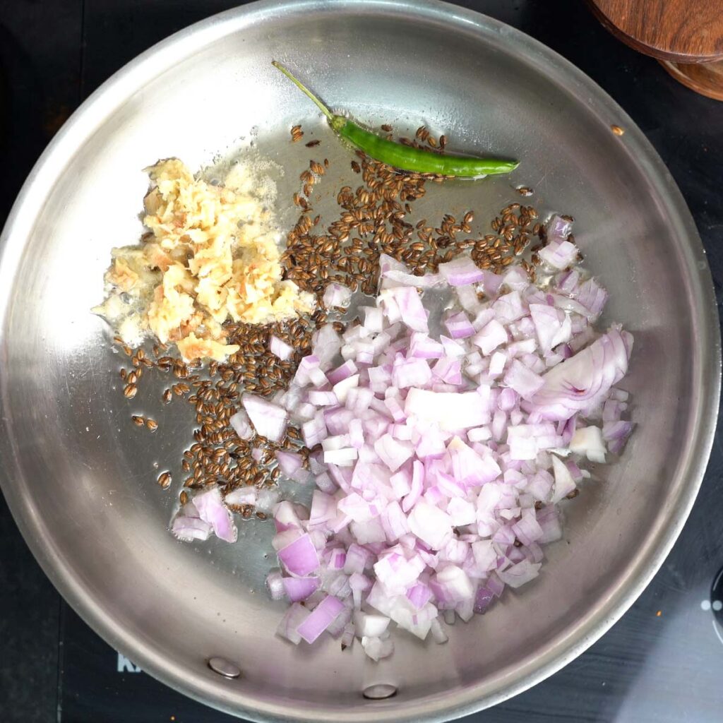 Adding crushed Ginger, garlic and chopped onion to a steel pan with ghee & Cumin