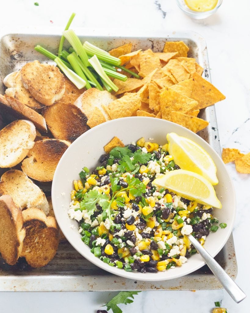 Black bean corn and feta dip in a grey bowl for a party with toasted bread, vegetables and chips