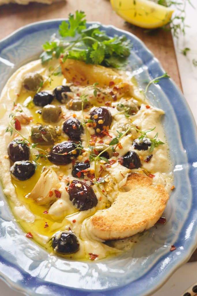 Hummus on a blue long platter with roasted olives, garlic, chilli flakes, lemon wedge and green herbs. Served with toasted bread