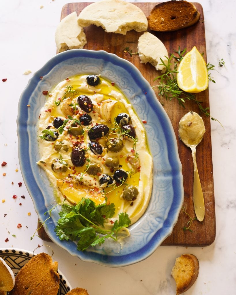 Hummus on a blue long platter with roasted olives, garlic, chilli flakes and green herbs