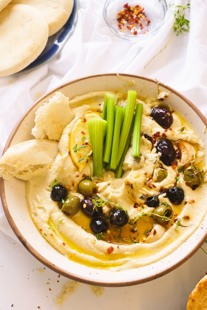 Hummus on a beige shallow bowl with roasted olives, garlic, chilli flakes, lemon wedge and green herbs. Served with warm pita bread and celery sticks