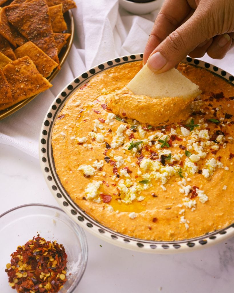 Dipping pita bread into a shallow plate of roasted red pepper feta dip.