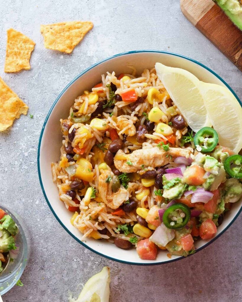 Instant Pot Burrito Bowl- Mexican Rice with Chicken & Beans in a white bowl with a green rim. Served along with wedges of lime, chips and a chunky guacomole