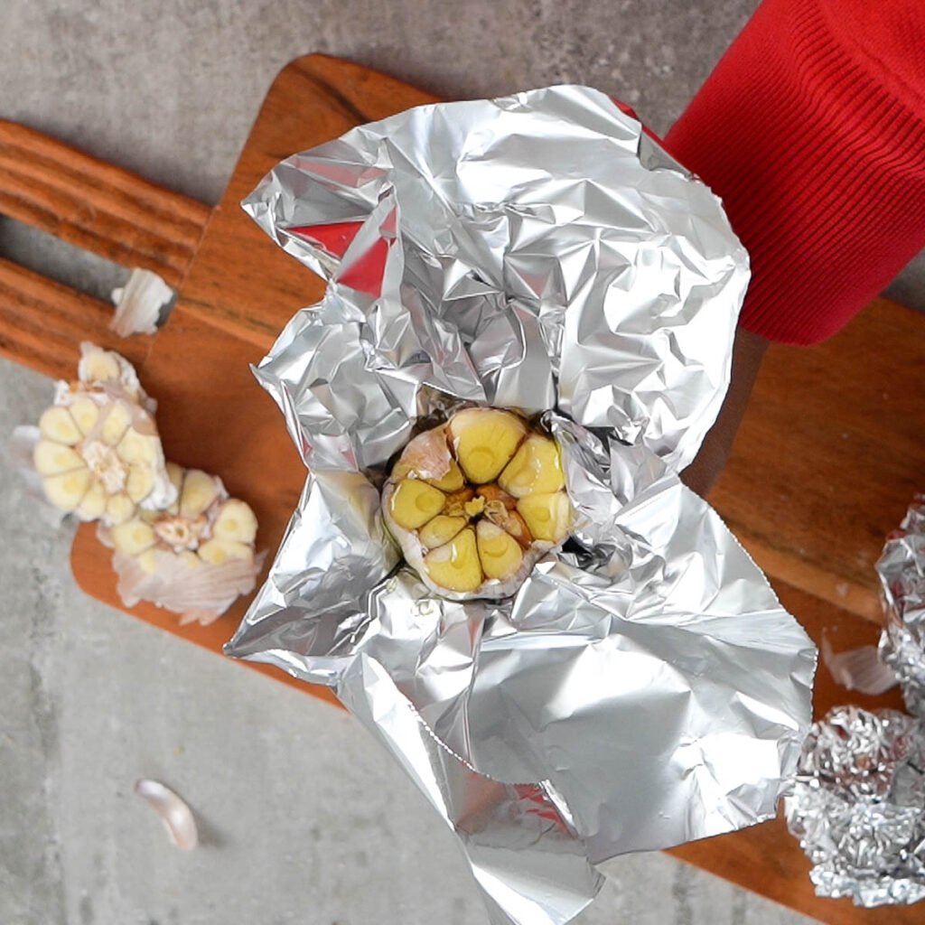 Aluminium foil wrapped garlic with olive oil and salt on a cutting board surrounded with more garlic