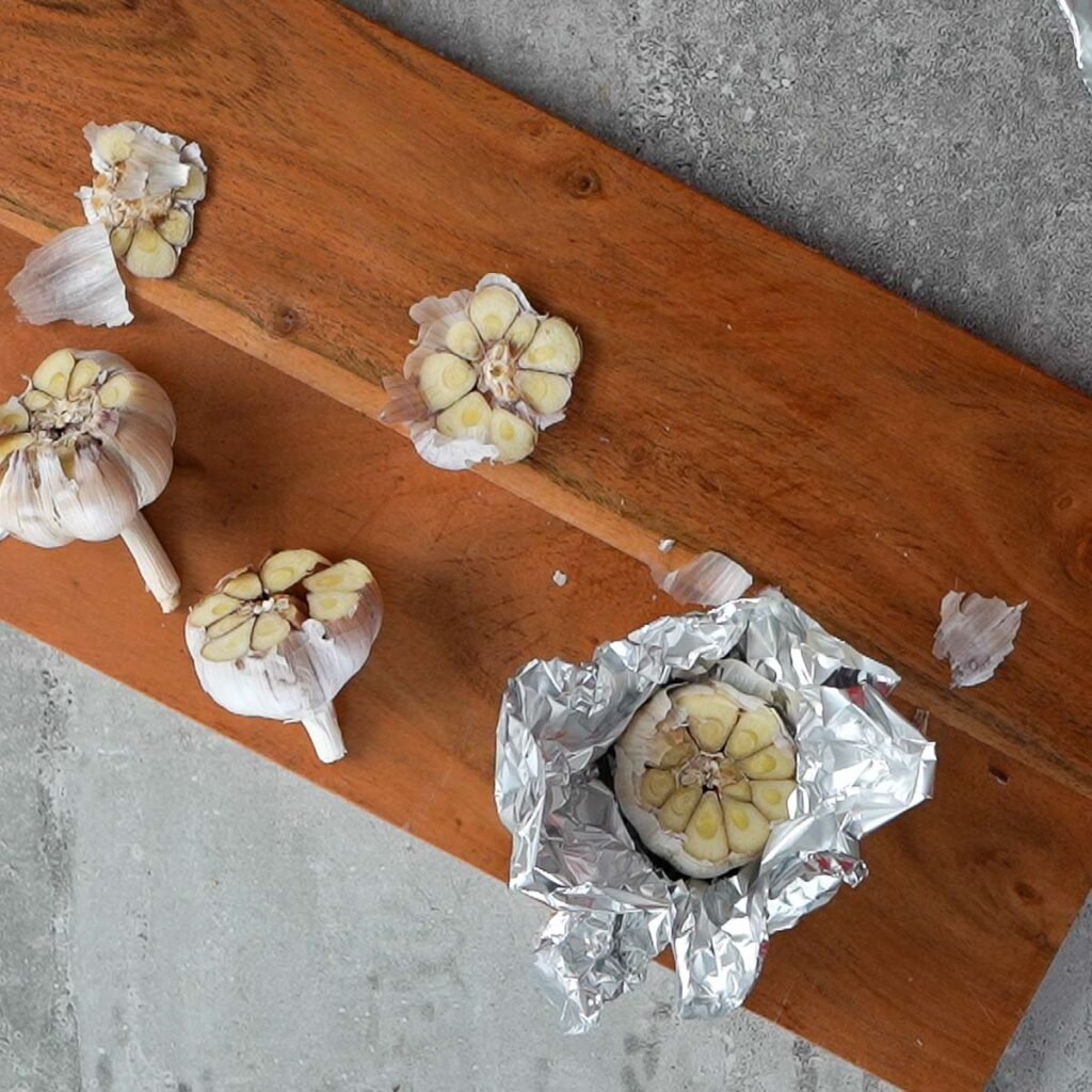 Garlic pods with their tops cut off. One garlic pod is inside aluminium foil on a wooden cutting board