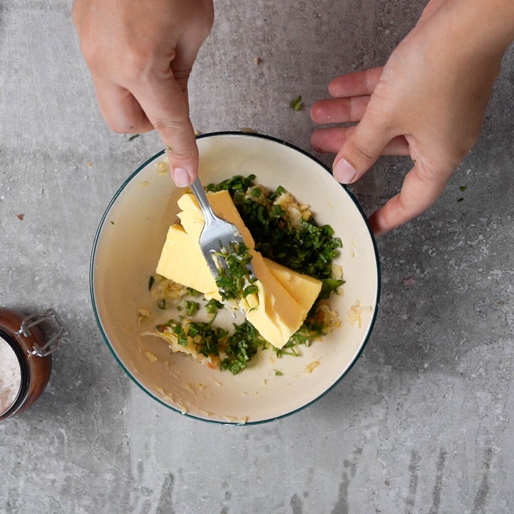 Butter being mashed into a bowl full of roasted garlic and herbs.