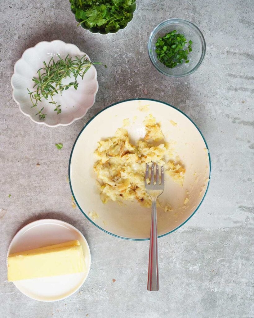 Ingredients needed to make Roasted Garlic Butter-mashed Roasted garlic in a white bowl, butter, herbs - rosemary, thyme, chives and cilantro in small bowls