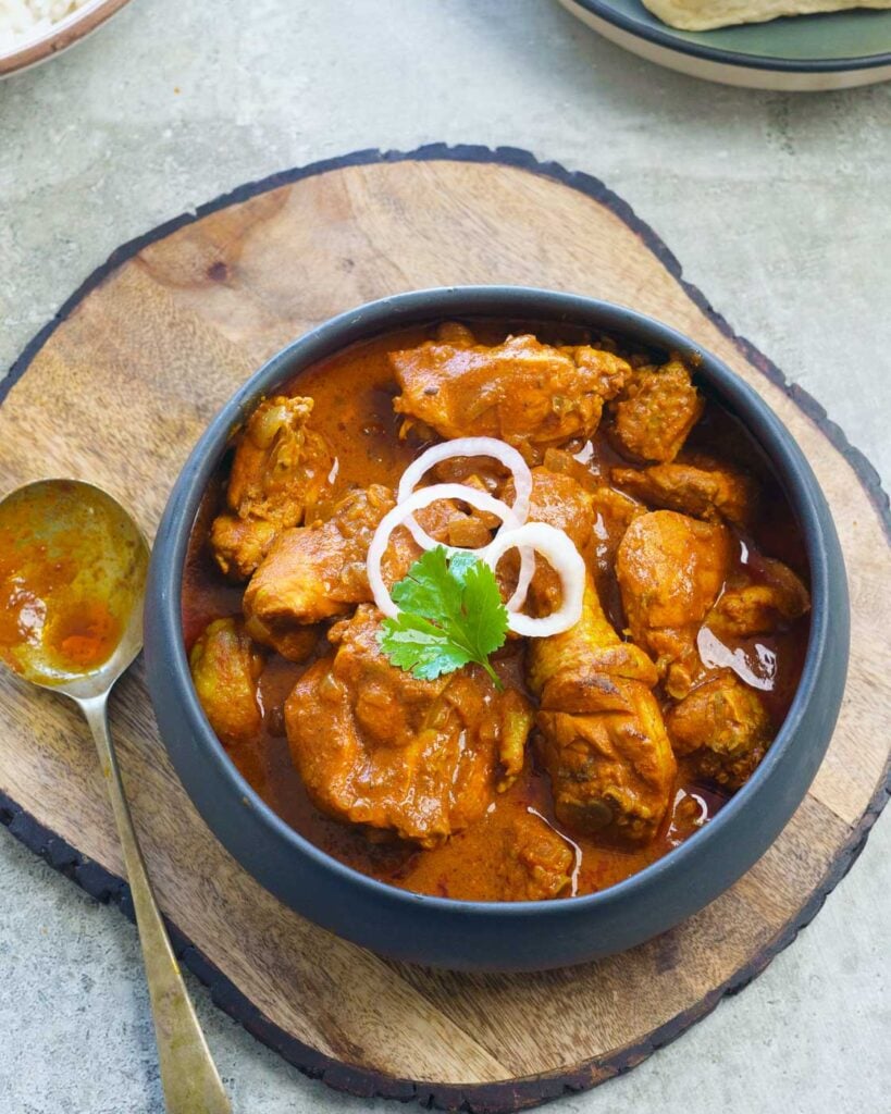 Chicken Vindaloo in a black bowl with 3 pieces of onion and a cilantro on top. Kept on a wooden trivet