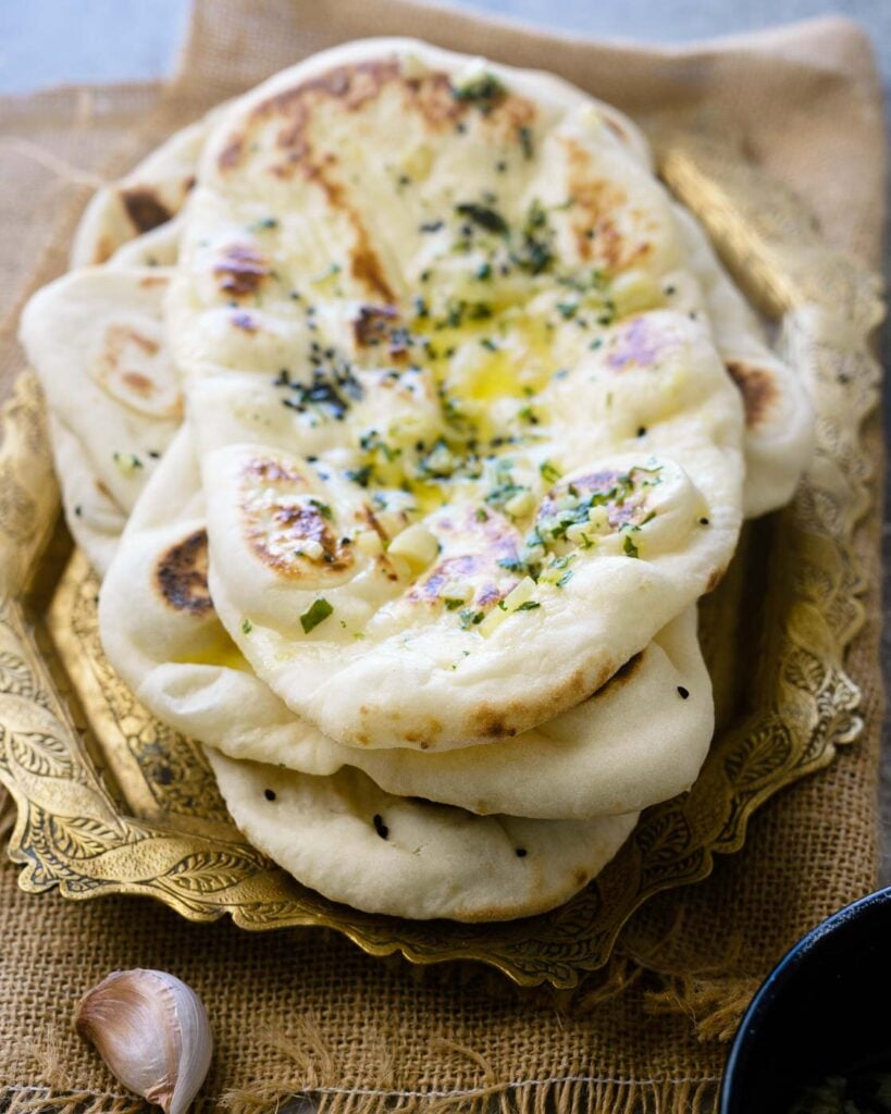 Soft pillowy butter garlic Naans stacked one on top of the other with garlic butter in a black bowl on the side