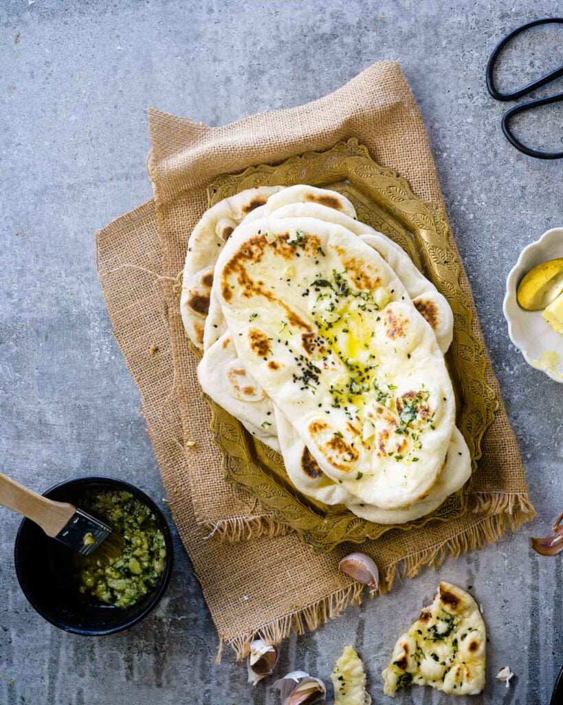Soft pillowy butter garlic Naans stacked one on top of the other with garlic butter in a black bowl on the side