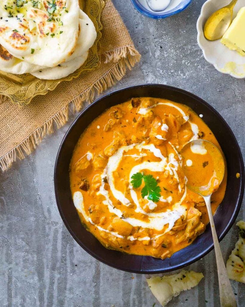 Murg Makhani or Butter Chicken in a black shallow bowl with a drizzle of cream, a few leaves of coriander and a naan on the side