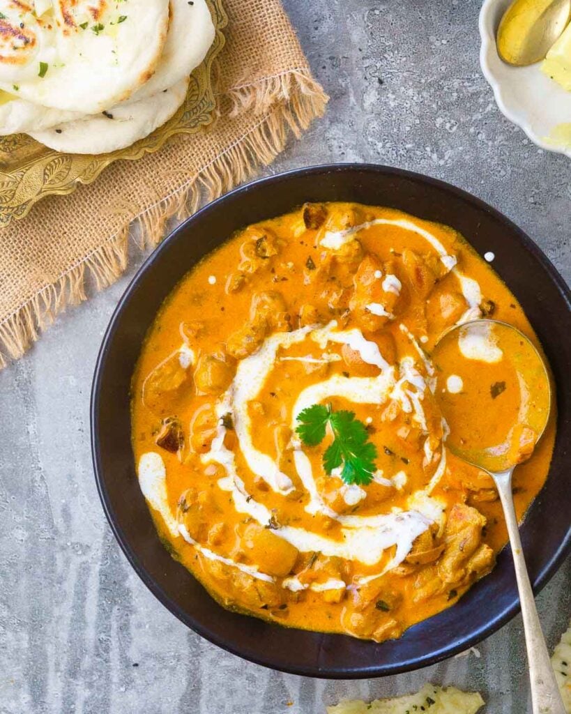 Murg Makhani or Butter Chicken in a black shallow bowl with a drizzle of cream, a few leaves of coriander and a naan on the side