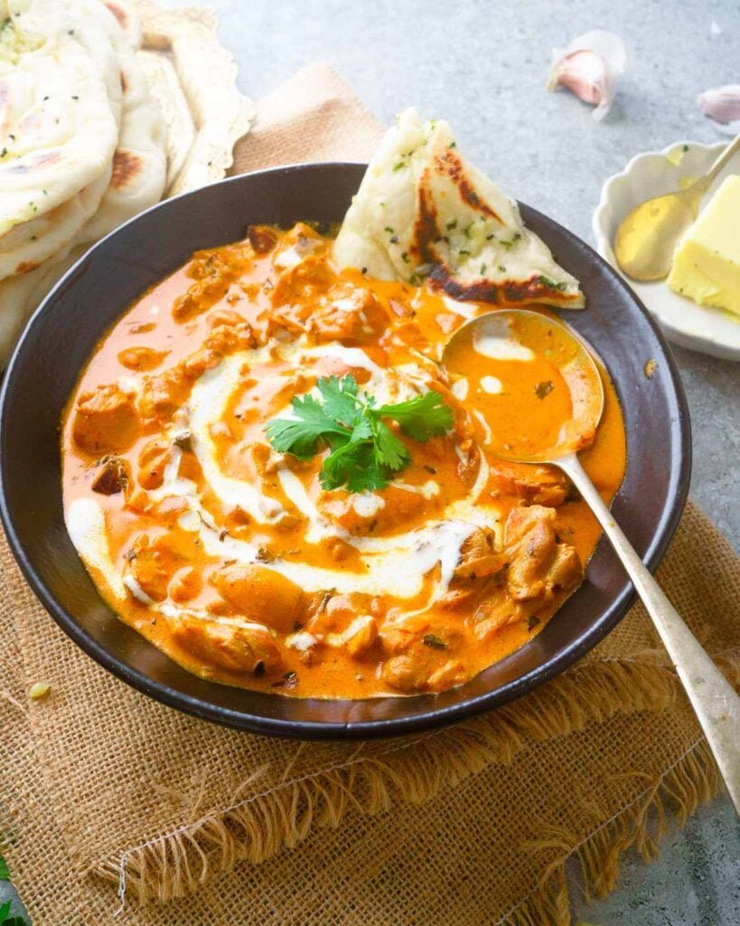 Murg Makhani or Butter Chicken in a black shallow bowl with a drizzle of cream, a few leaves of coriander and a naan on the side