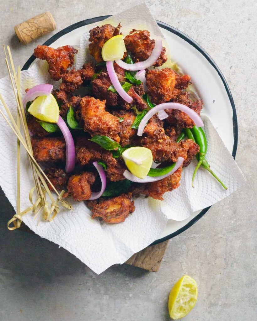 Chicken 65- indian spicy fried chicken on a white enameled plate with slices of onion, wedges of lime, fried curry leaves and tooth picks on a grey background