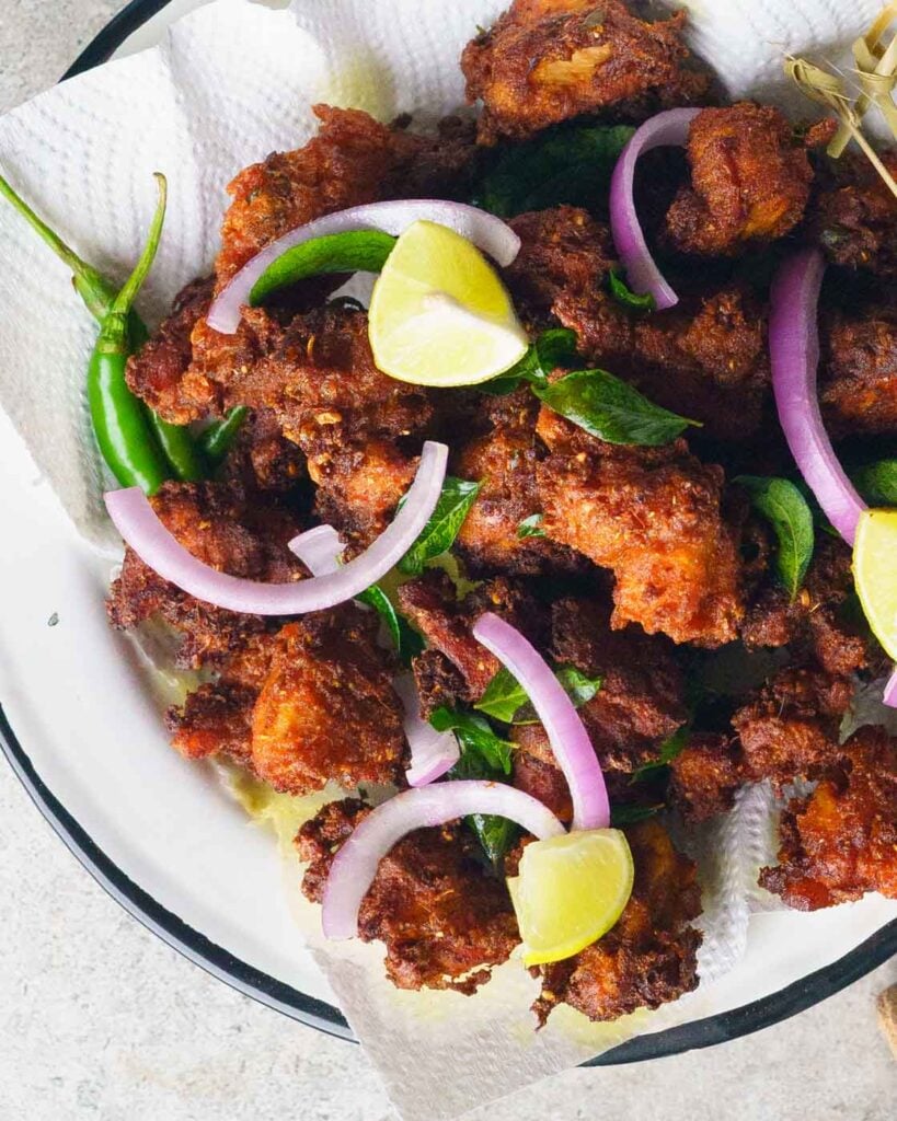 Chicken 65- indian spicy fried chicken on a white enameled plate with slices of onion, wedges of lime, fried curry leaves and tooth picks on a grey background