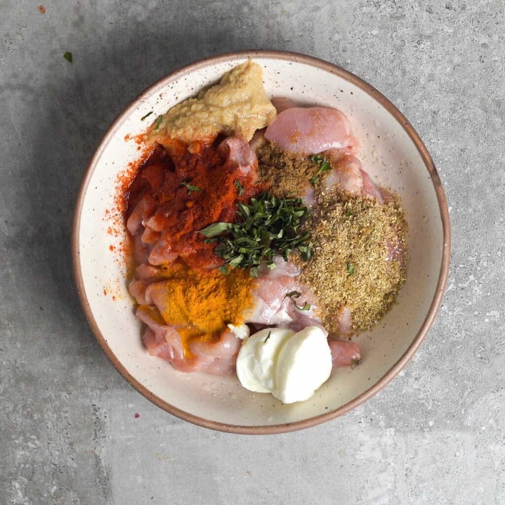 cut pieces of chicken thigh in a white bowl along with chili, coriander, turmeric, salt, ginger garlic paste, yogurt ,salt and sliced curry leaves