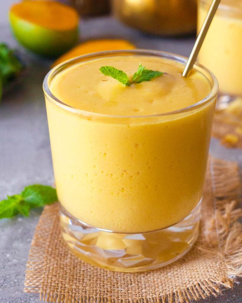 Mango Lassi in a glass with a sprig of mint on top with a golden stirer