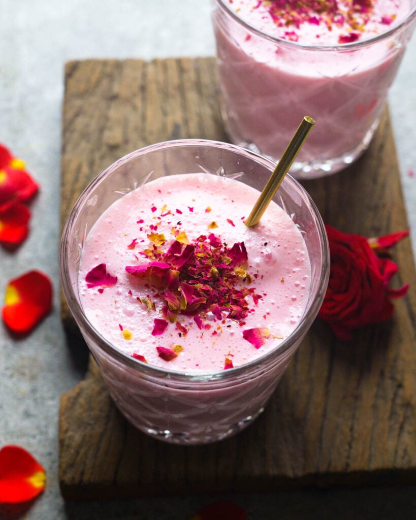 Rose Lassi in two glasses kept on a wooden plank with crushed rose petals, pistachios and a golden stirrer inside.