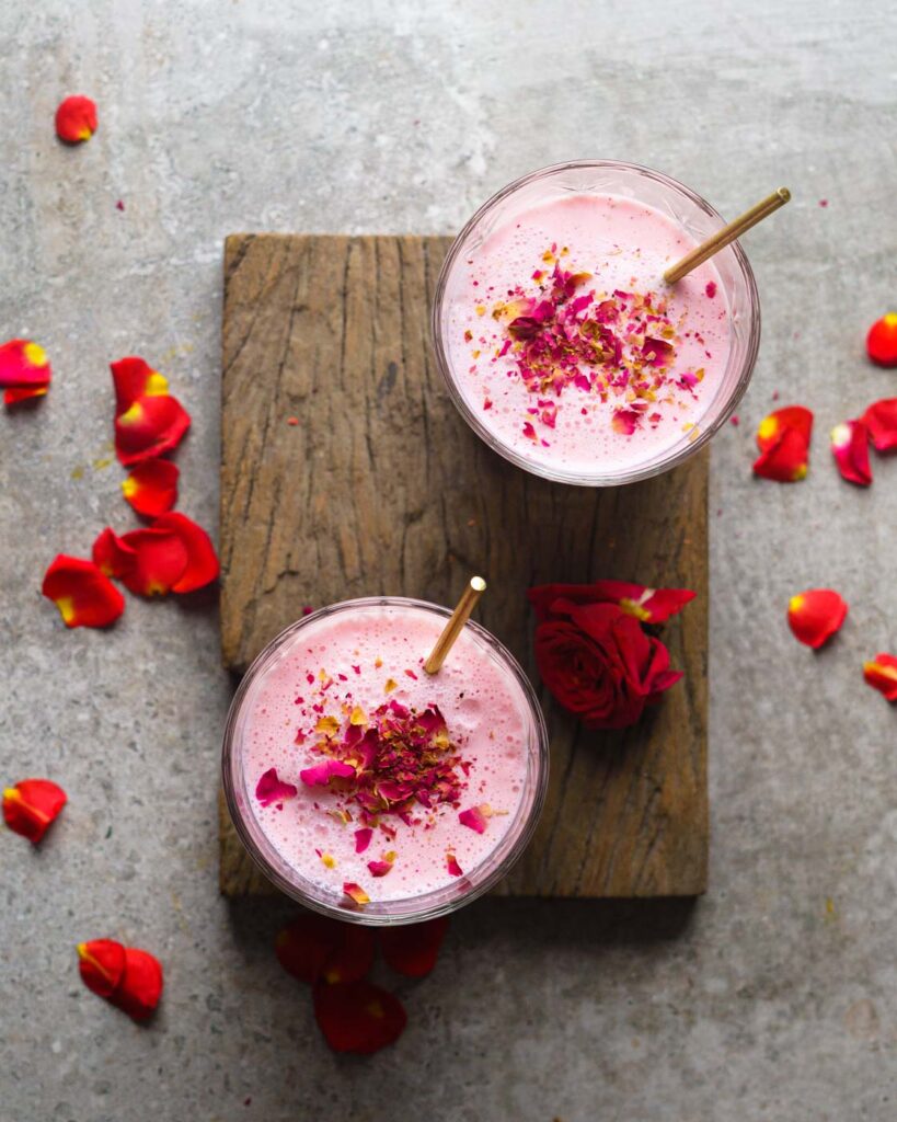 Rose Lassi in two glasses kept on a wooden plank with crushed rose petals, pistachios and a golden stirrer inside.