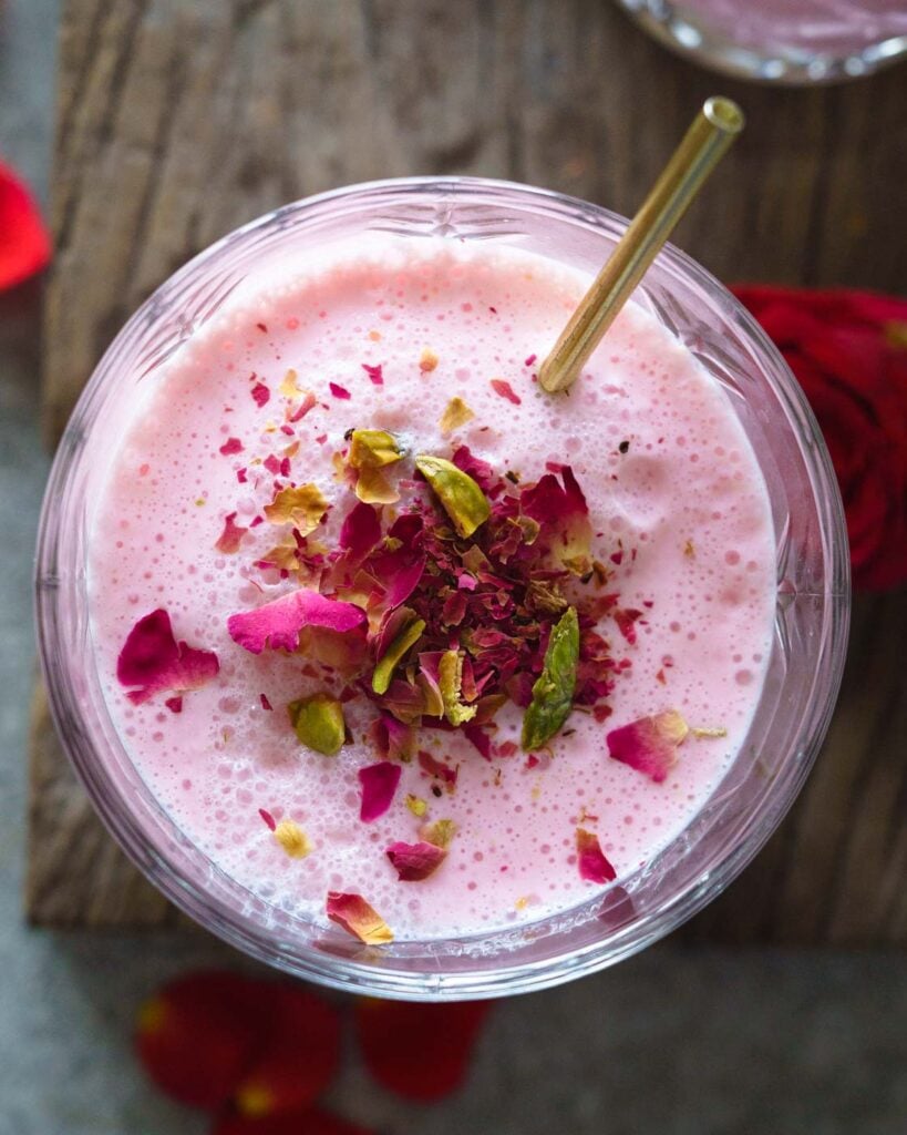 Rose Lassi in a glass kept on a wooden plank with crushed rose petals, pistachios and a golden stirrer inside.