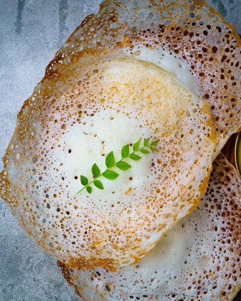 Three Appams or hoppers on a wooden plate along with one sprig of curry leaf on the middle appam