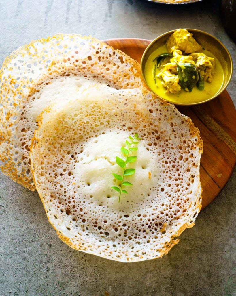 Three Appams or hoppers on a wooden plate along with chicken curry and one sprig of curry leaf on the middle appam