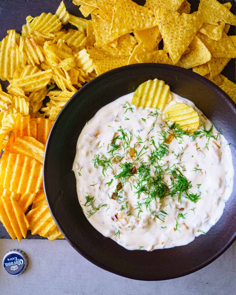 Crinkled chips and nachos around a bowl of yogurt caramalised onion dip with dill leaves in a black bowl