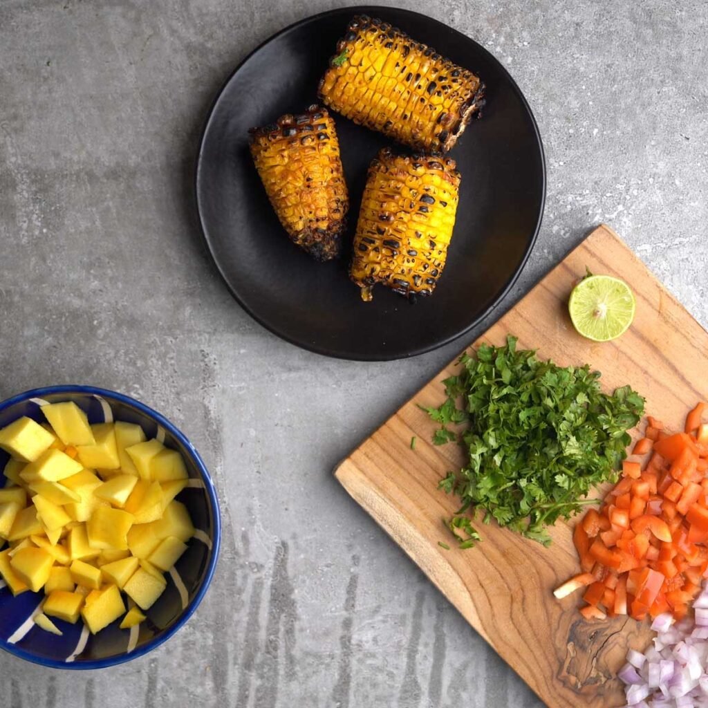 Grilled corn on the cob in a black plate, with chopped up cilantro, bell pepper. lime and onion, along with a blue bowl of mango