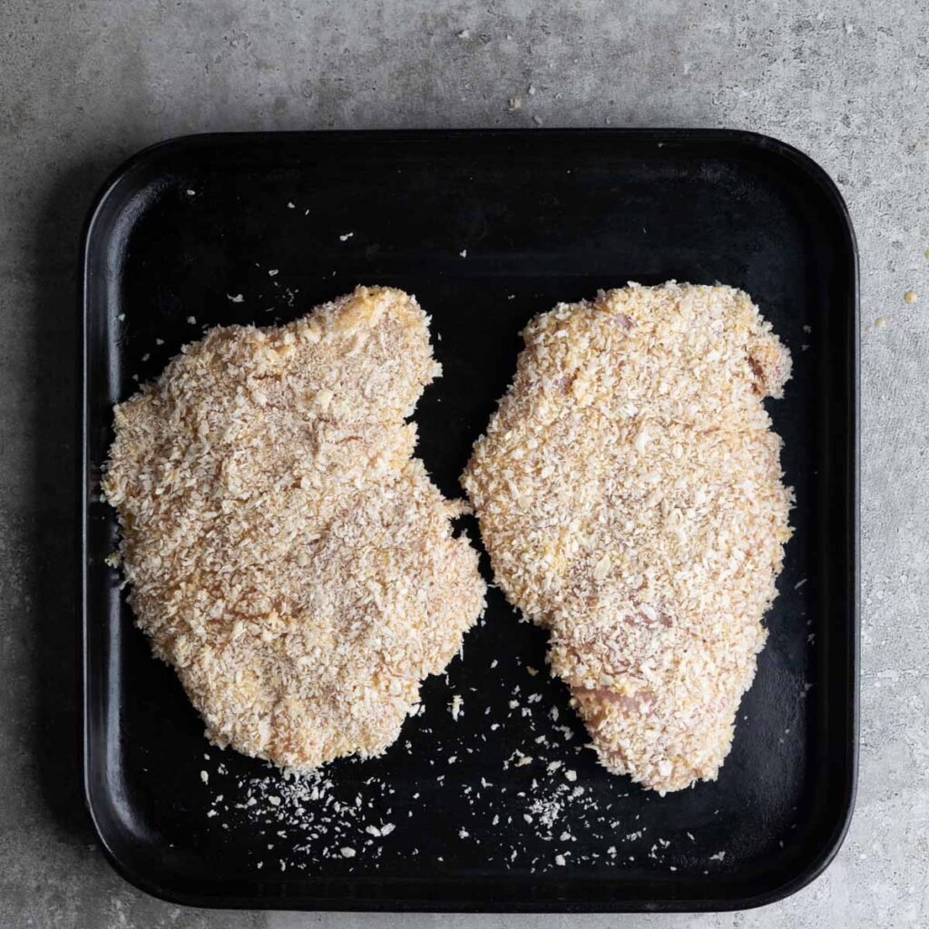 panko bread crumbs coated chicken breast steaks on a black baking tray