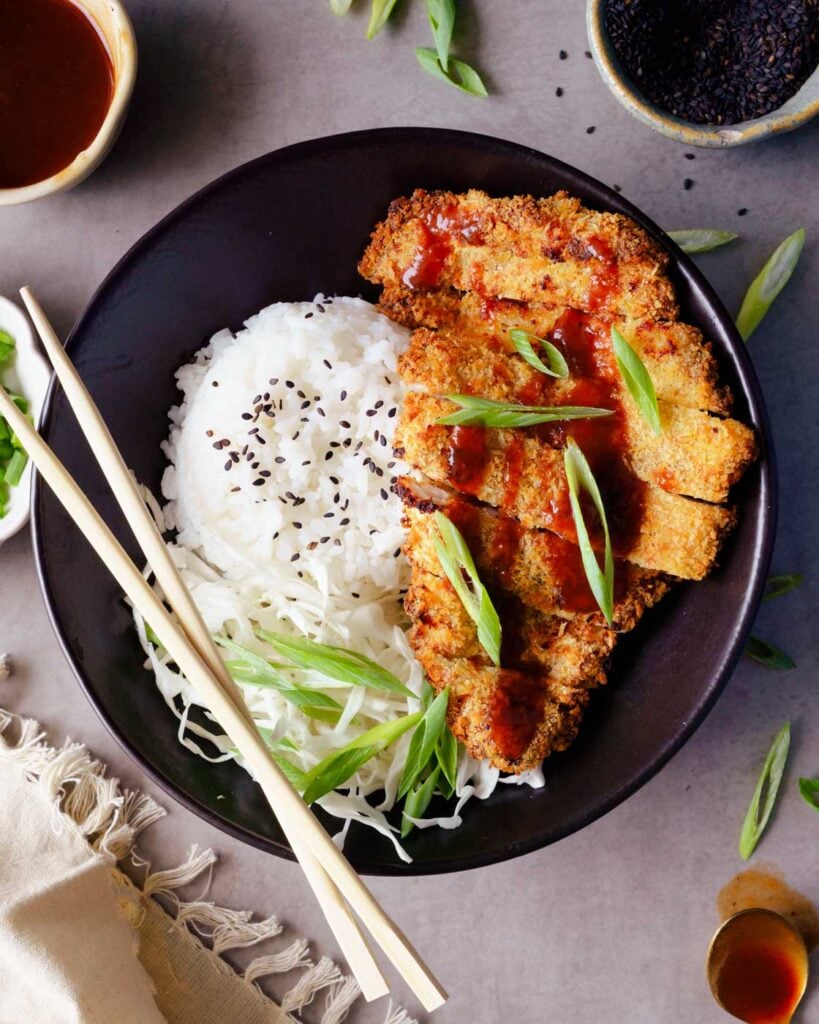 Chicken Katsu served in a black bowl with steamed rice, a generous drizzle of tonkatsu sauce, spring onions & shredded Cabbage.