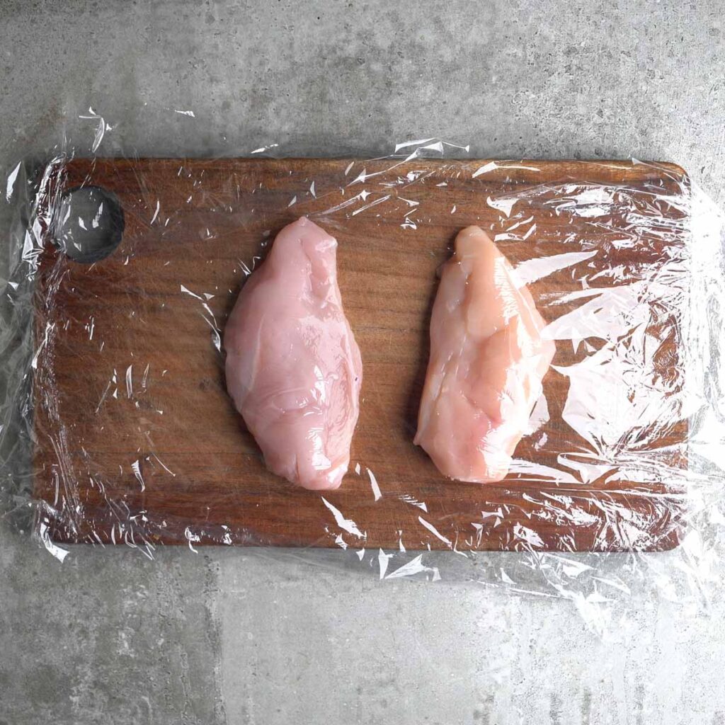 2 chicken breasts on a cutting board lined with plastic wrap