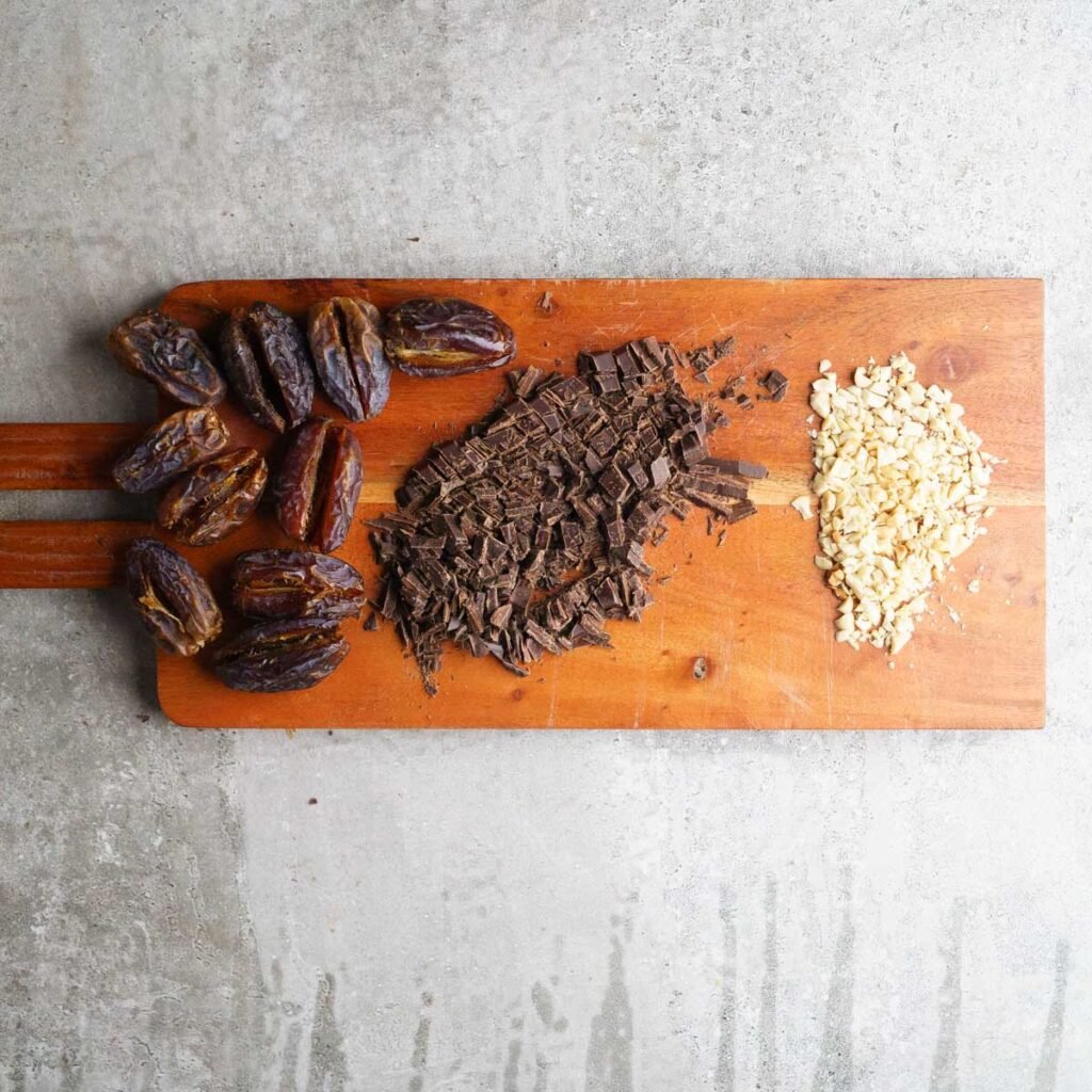 Chopped chocolate, peanuts and slit medjool dates on a long wooden chopping board