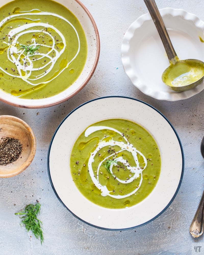 Bright green Zucchini Soup in a white soup bowl with a drizzle of olive oil and cream.On the side you can see another bowl with soup and a ladle that was used to take the soup