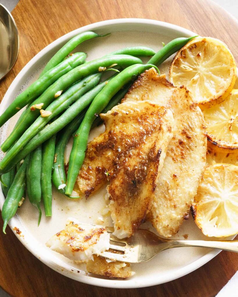 Flaky tilapia fish, served with french beans on a white plate along with slices of lemon.