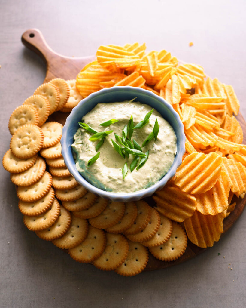 Green onion dip served in a blue bowl on a wooden round platter, along with ritz crackers and potato chips.