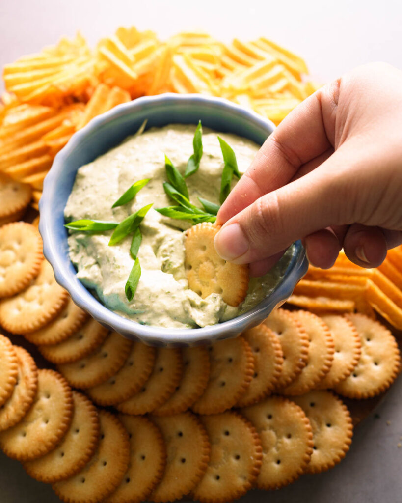 Green onion dip served in a blue bowl, along with ritz crackers and potato chips. A ritz cracker being dipped inside.