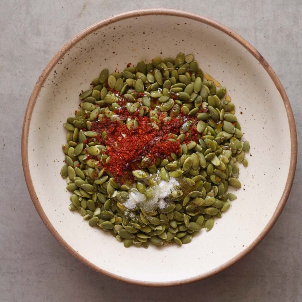 pumpkin seeds or pepitas in a white bowl with brown rim topped with ground chili, salt, oil and maple syrup