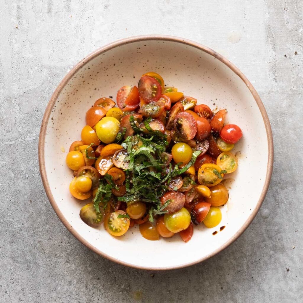 Cherry tomatoes quartered in a white bowl along with shredded basil leaves, olive oil and salt