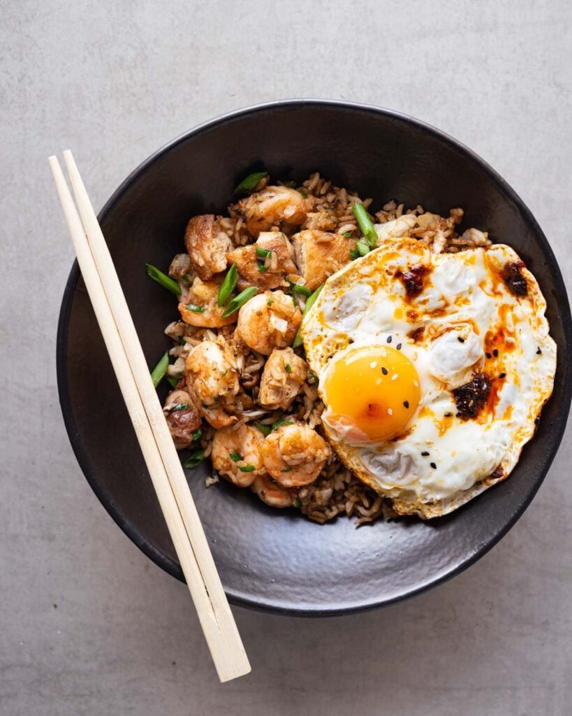 Shrimp and chicken fried rice in a black bowl, garnished with green onions, a fried egg with chili oil, black and white sesame seeds.